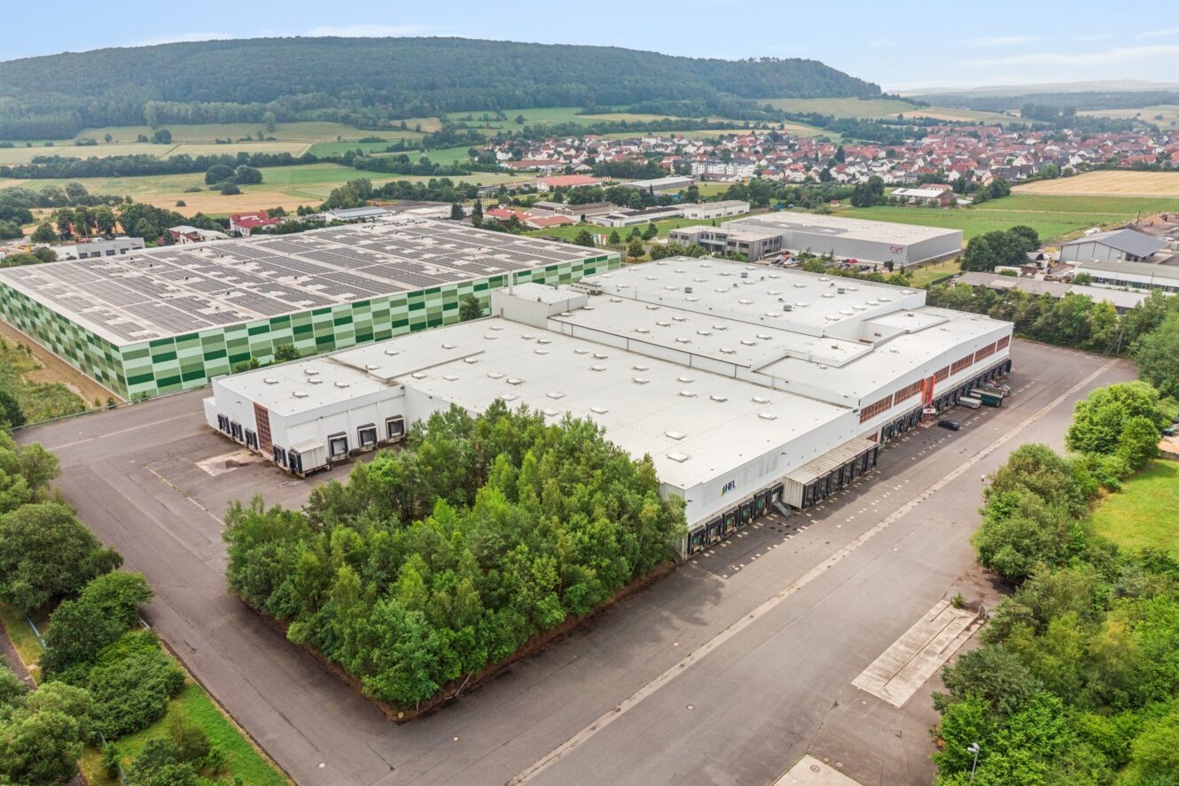 Luftaufnahme eines großen, von Bäumen umgebenen Industrie- und Lagerkomplexes mit nahegelegenen Gebäuden, offenen Parkplätzen und einer im Hintergrund sichtbaren Stadt.