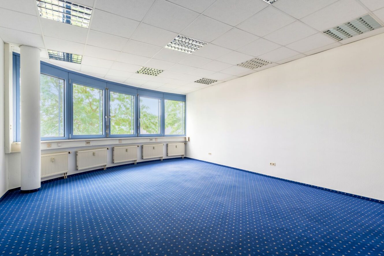 Ein leerer Büroraum mit blauem Teppichboden, weißen Wänden, großen, geschwungenen Fenstern und Deckenleuchten. Durch die Fenster sind draußen Bäume zu sehen.