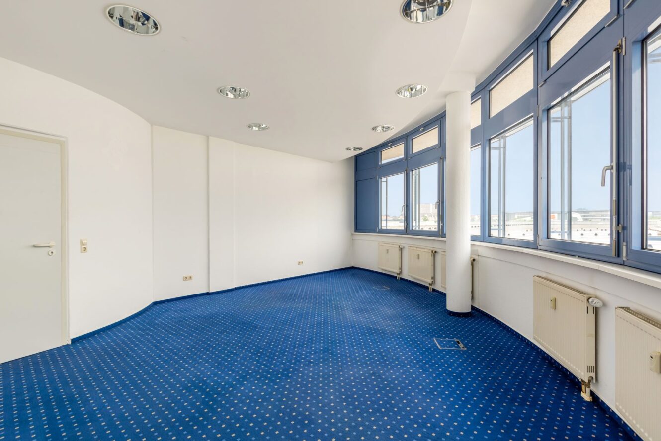 Ein leerer Raum mit einem blau gepunkteten Teppich, weißen Wänden, einer gewölbten Decke, mehreren Deckenleuchten, großen Fenstern und Heizkörpern entlang der Wand.