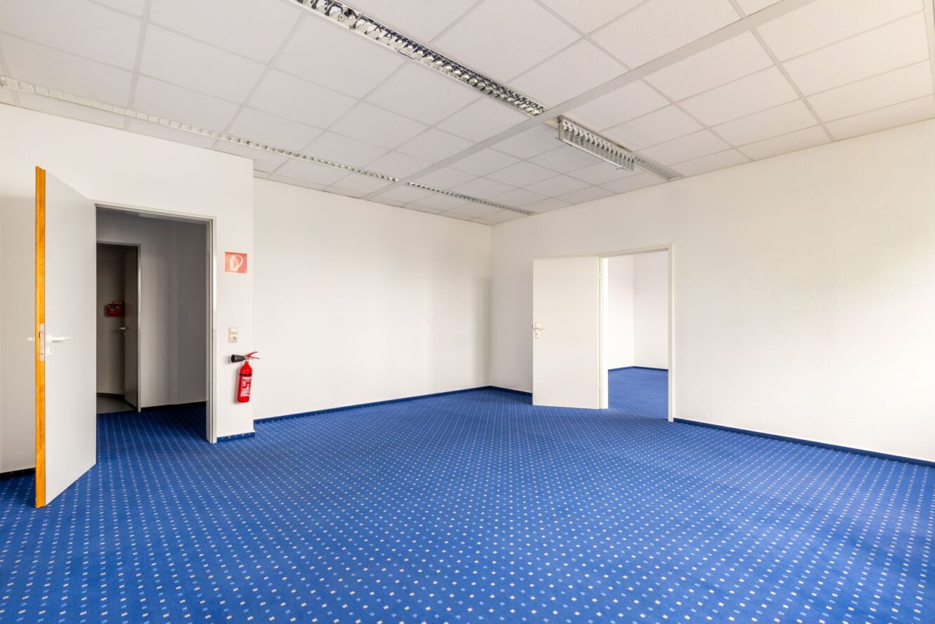 Ein leeres Büro mit blauem Teppichboden, weißen Wänden, zwei offenen Türen, Deckenleuchten und einem an der Wand montierten Feuerlöscher in der Nähe des Eingangs.