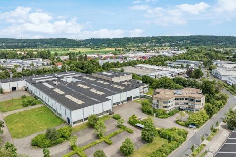 Luftaufnahme eines großen Industriekomplexes mit mehreren Lagerhallen und einem angrenzenden Bürogebäude, umgeben von Grünflächen und Parkplätzen. In der Nähe sind Straßen und in der Ferne Hügel zu sehen.