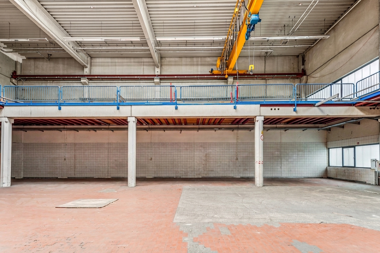 Leere Industriehalle mit gefliesten Wänden, Betonsäulen und einem blauen Laufkran. Der Boden ist teilweise mit roten und grauen Fliesen ausgelegt. Entlang der Rückwand verläuft ein erhöhter Laufsteg.