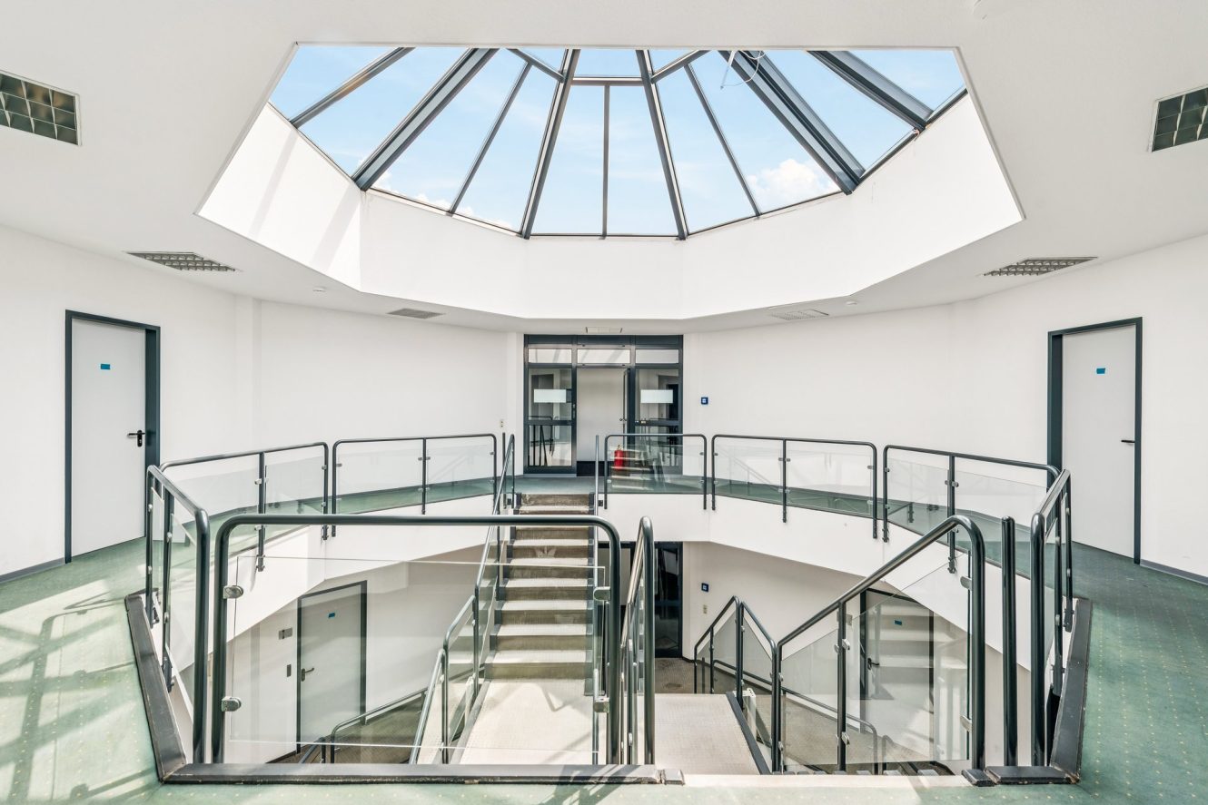 Ein moderner Innenraum mit einem Glasoberlicht, grünem Teppichboden, weißen Wänden und einer zentralen Treppe, die von Glasgeländern umgeben ist. Im Obergeschoss reihen sich mehrere Türen aneinander.