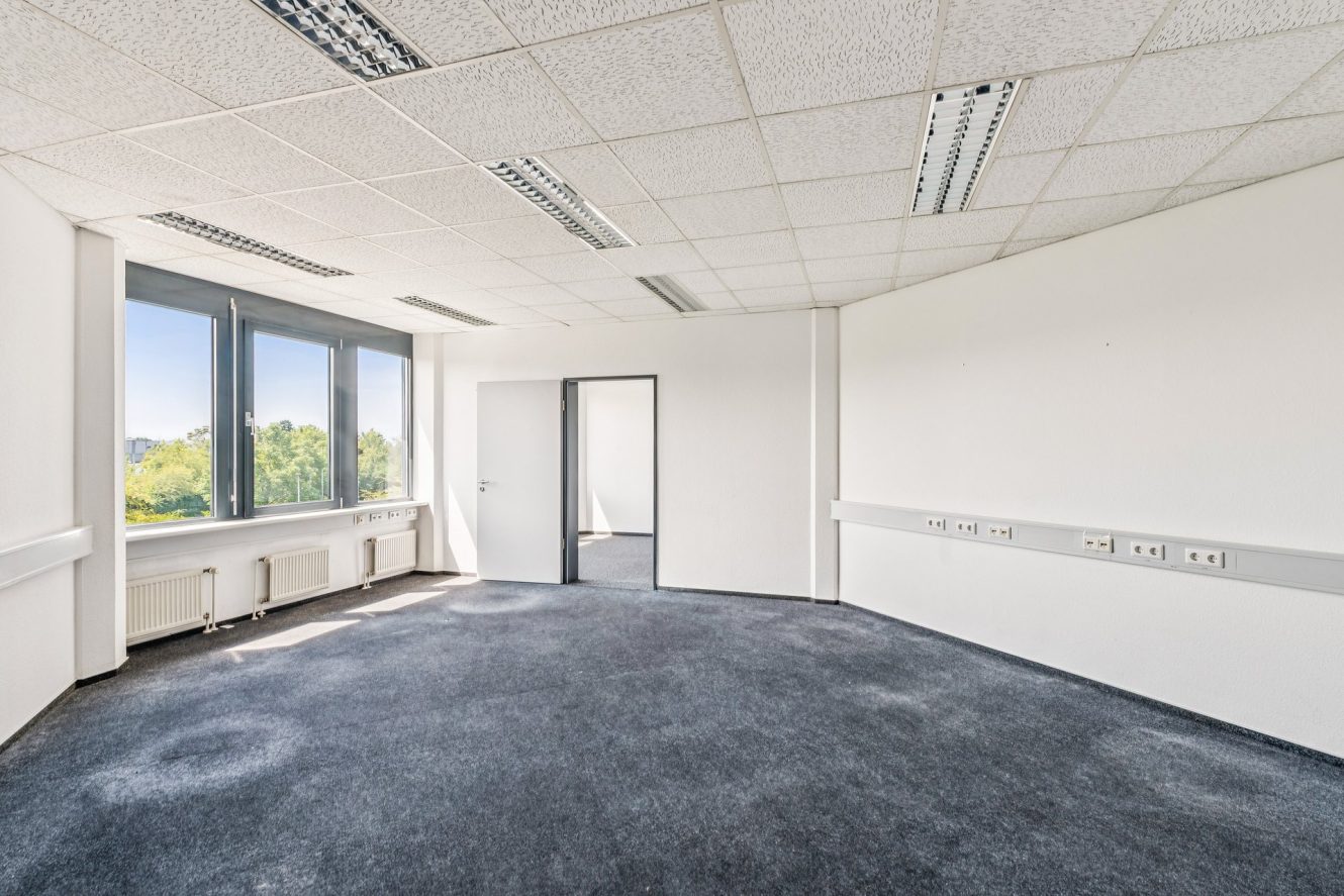 Leerer Büroraum mit Teppichboden, großen Fenstern, weißen Wänden, Deckenleuchten und Steckdosen entlang einer Wand. Eine Tür führt in einen angrenzenden Raum.