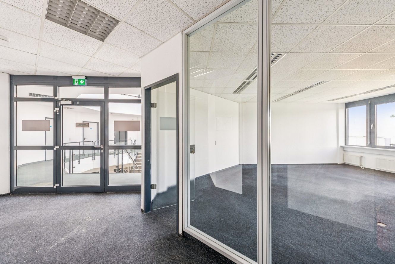 Leere, moderne Büroräume mit Glaswänden, schwarzem Teppichboden, weißen Decken, Leuchtstoffröhren und großen Fenstern; Doppeltüren führen in einen Flur mit einer Treppe und einem Notausgangsschild über dem Kopf.