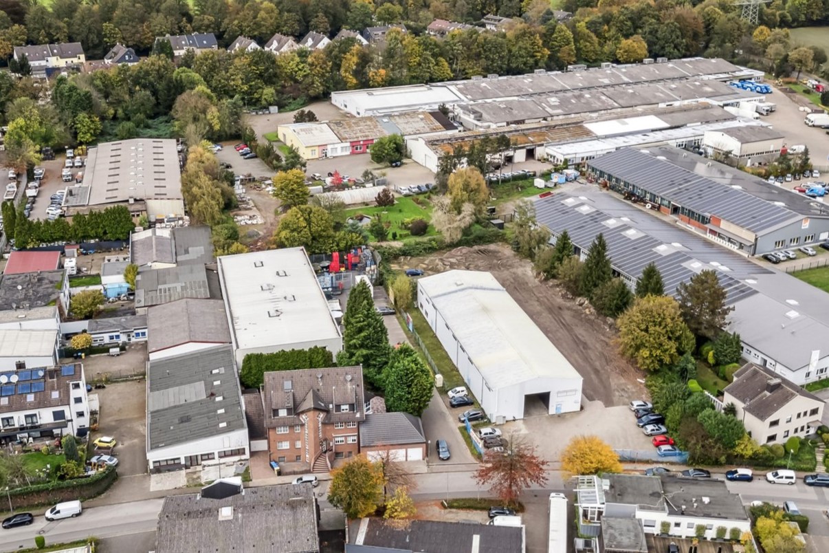 Luftaufnahme eines Industriegebiets mit zahlreichen Lagerhallen, Geschäftsgebäuden, geparkten Fahrzeugen und umliegenden Bäumen in der Nähe eines Wohngebiets.