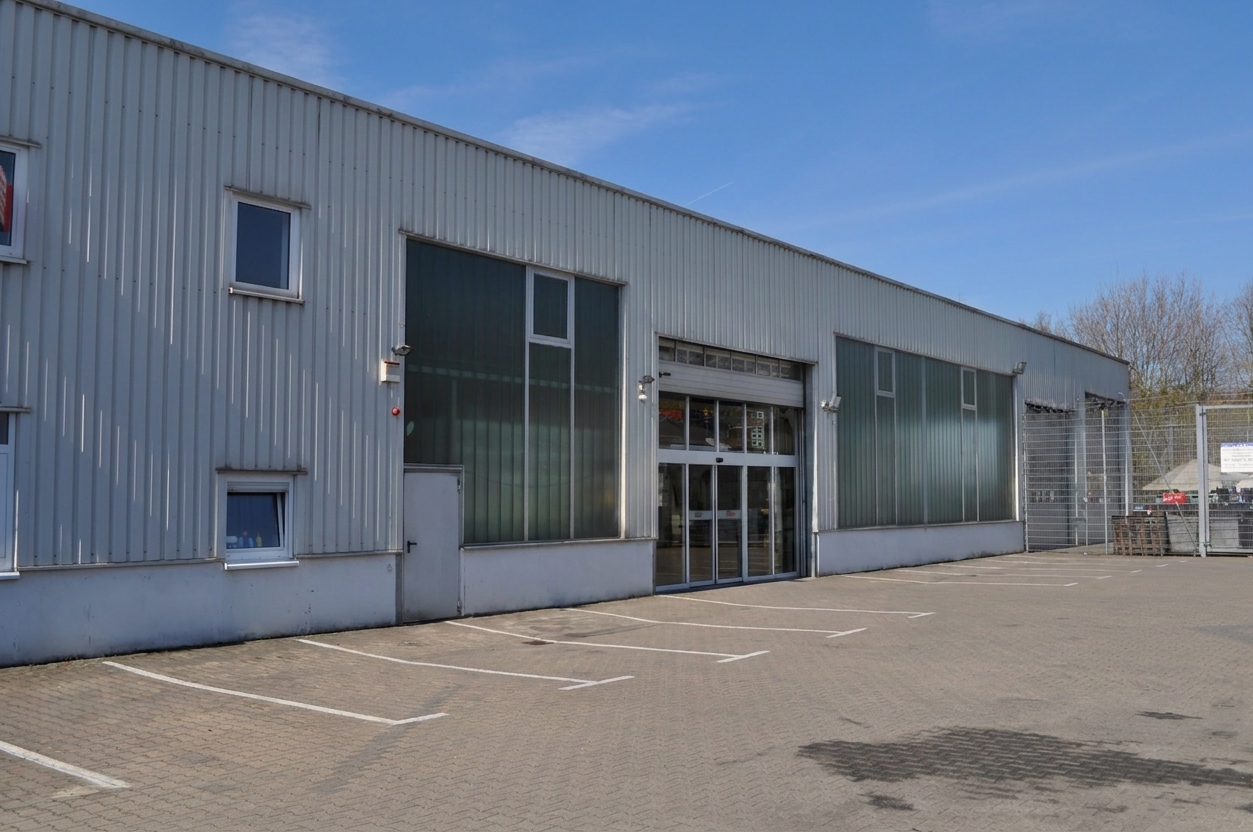Inmitten eines leeren Parkplatzes steht unter strahlend blauem Himmel ein großes Industriegebäude mit Metallverkleidung, Glastüren und mehreren Fenstern.