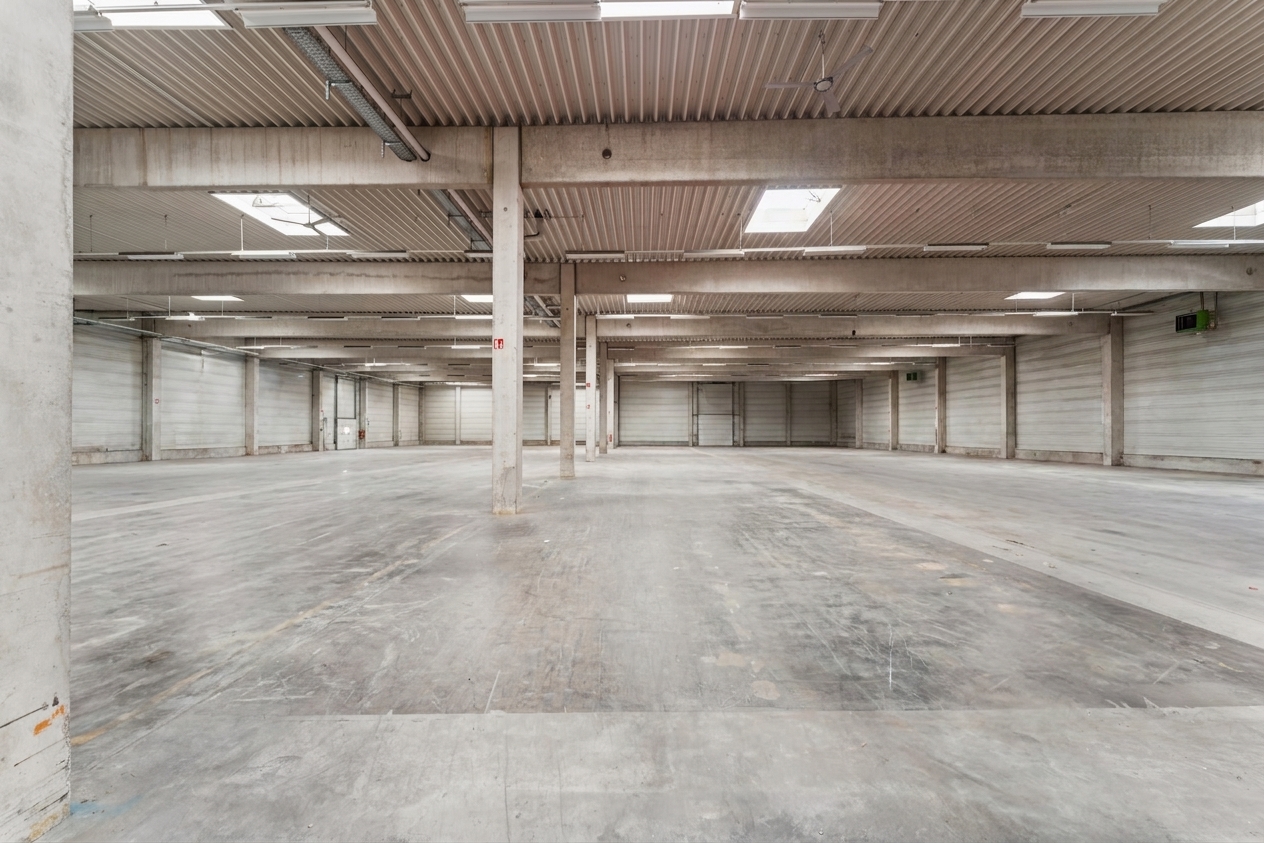 Leere Industriehalle mit Betonböden, hohen Decken, Stützpfeilern und Oberlichtern. Der Raum ist durch Tageslicht und Leuchtstoffröhren gut ausgeleuchtet.