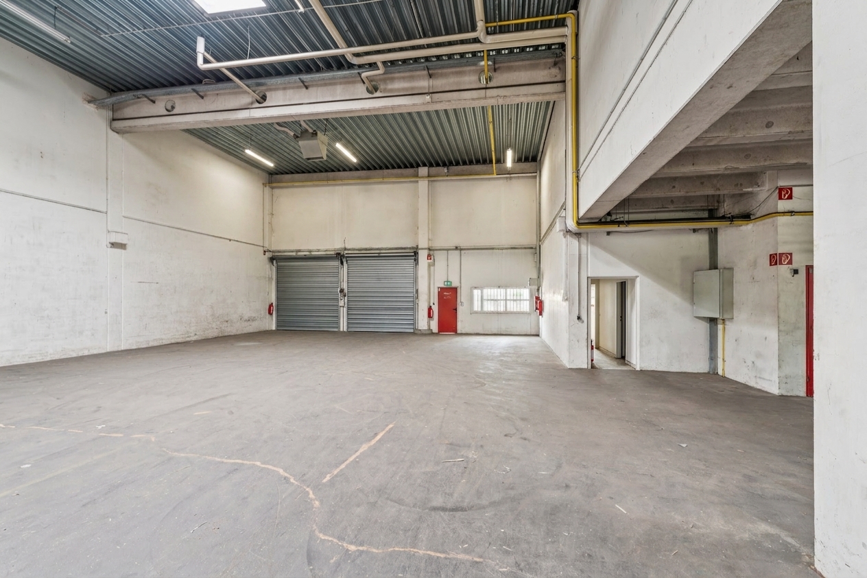 Innenansicht einer leeren Industriehalle mit Betonboden, hoher Decke, großem Rolltor, Leuchtstoffröhren und weißen Wänden. Notausgänge und Feuerlöscher sind zu sehen.