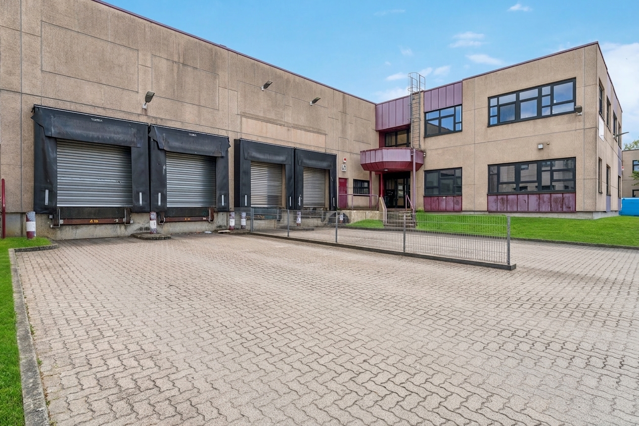 Ein großes Industriegebäude mit drei Laderampen, geschlossenen Metalltoren, einer Treppe, Fenstern und einem gepflasterten Vorplatz.