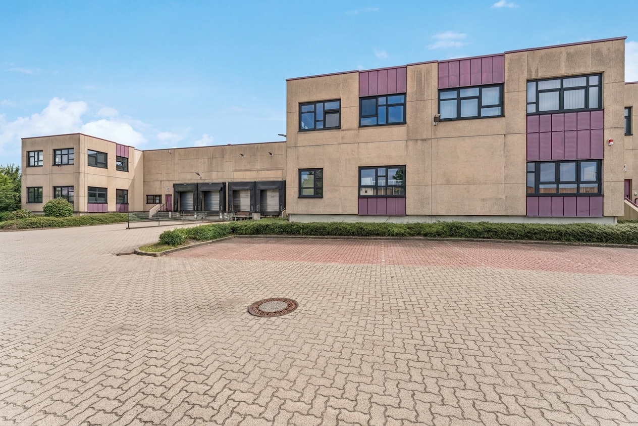 Ein großes Industriegebäude mit beigen Wänden, violetten Verkleidungen, zahlreichen Fenstern und drei Laderampen, gelegen auf einem weitläufigen, gepflasterten Grundstück unter strahlend blauem Himmel.