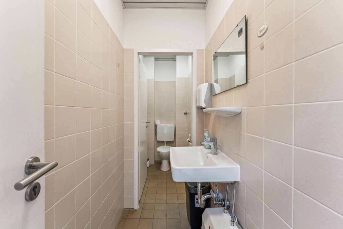 Ein kleines Badezimmer mit beige gefliesten Wänden, einem weißen Waschbecken unter einem Spiegel, einem Seifenspender am Waschbecken und einer Toilette, die hinten steht.