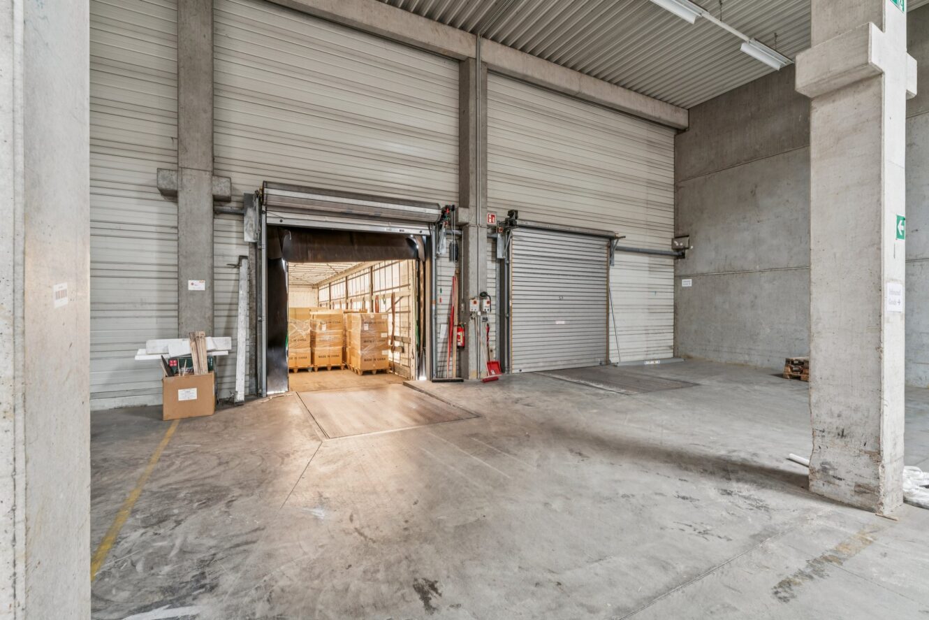 Innenansicht einer Laderampe in einer Industriehalle mit geöffneten Rolltoren, Paletten sowie Betonböden und -wänden.