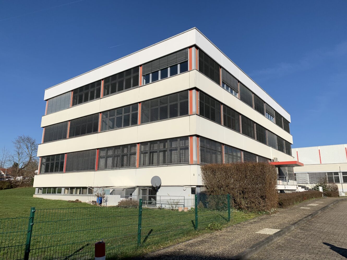 Unter einem strahlend blauen Himmel steht ein modernes, vierstöckiges Bürogebäude mit großen Fenstern und roten vertikalen Akzenten neben einer umzäunten Rasenfläche.