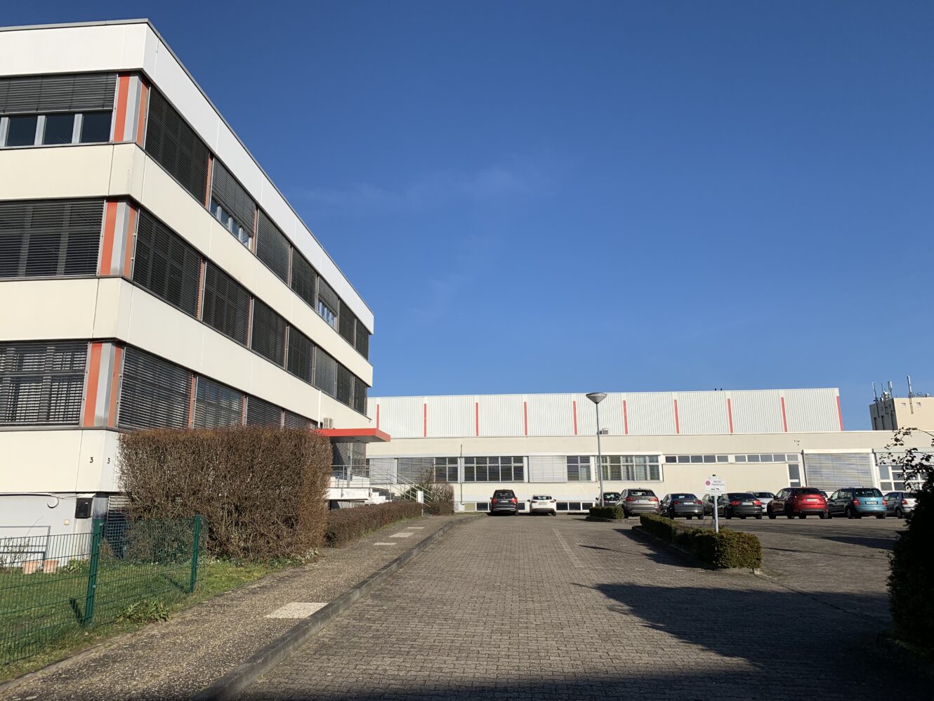 Moderne Büro- und Industriegebäude mit großen Fenstern, einer gepflasterten Zufahrt, geparkten Autos und einem strahlend blauen Himmel.