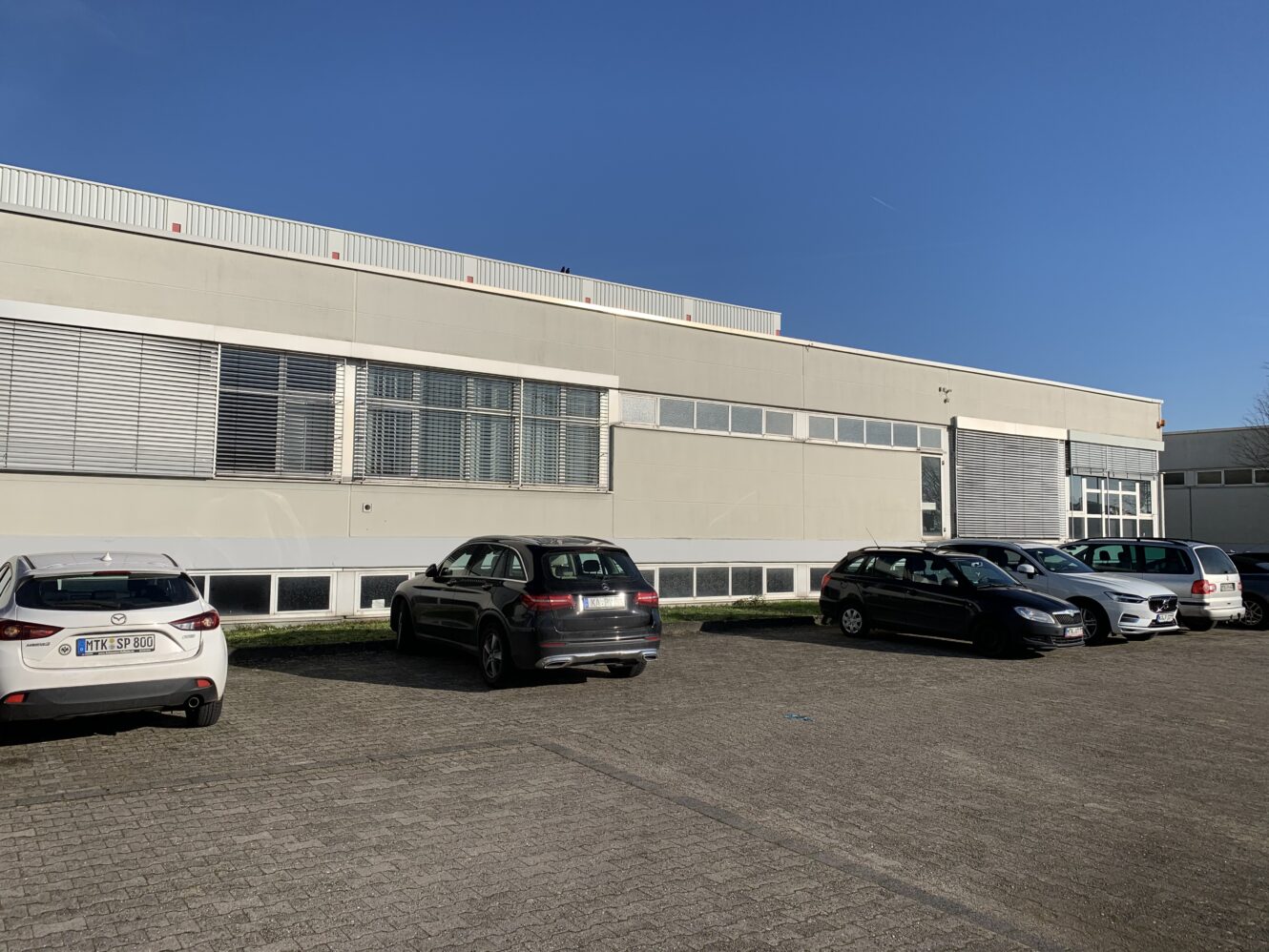 An einem sonnigen Tag steht eine Reihe von Autos vor einem großen, hellen Industriegebäude mit vielen Fenstern.
