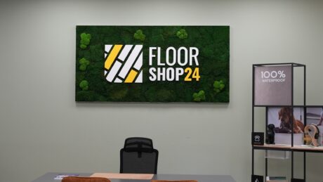 Bürowand mit einem „FLOOR SHOP 24“-Schild und -Logo über einem Schreibtisch, daneben ein Regal, auf dessen rechter Seite ein Etikett mit der Aufschrift „100 % WASSERDICHT“ zu sehen ist.