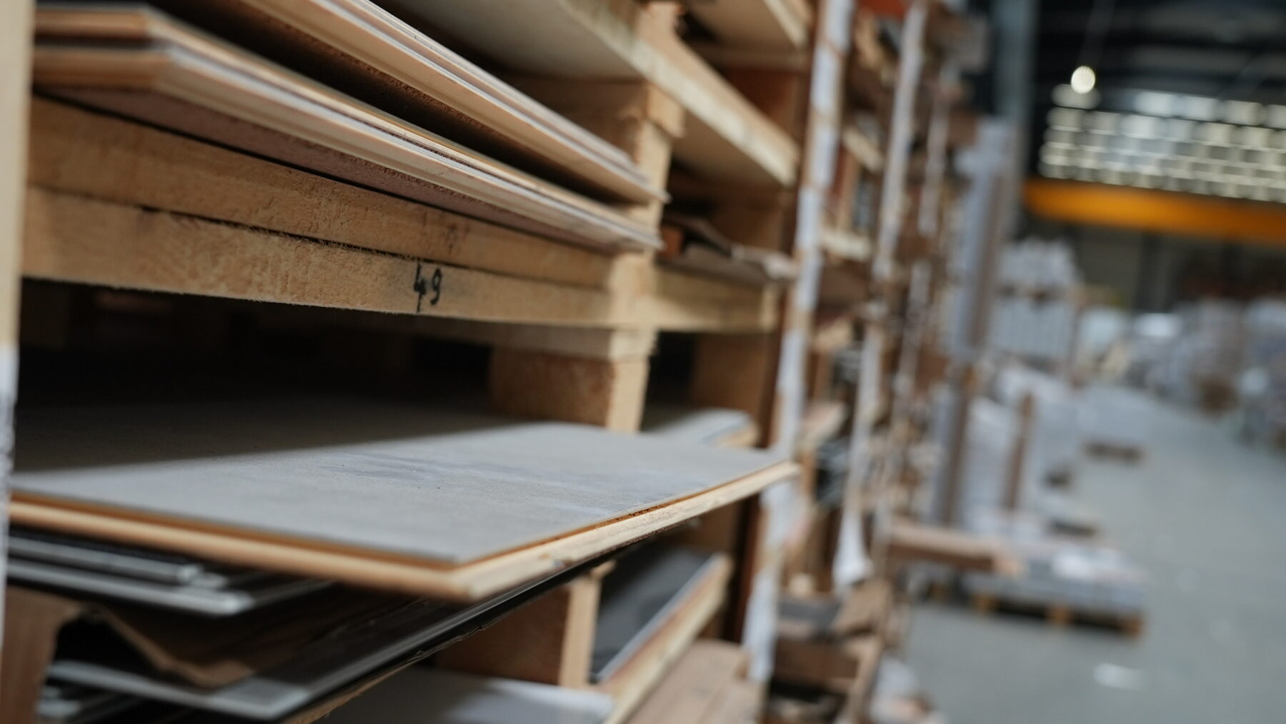 Nahaufnahme von gestapelten Holzpaletten, auf denen große Fliesen oder Bretter in einem Lager liegen; im Hintergrund sind weitere Regale und Industriebeleuchtung zu sehen.