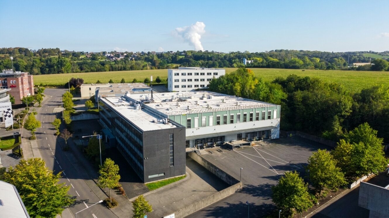 Luftaufnahme eines modernen Industriegebäudes mit weißem Dach, umgeben von Bäumen, leeren Parkplätzen und Feldern im Hintergrund unter einem strahlend blauen Himmel.