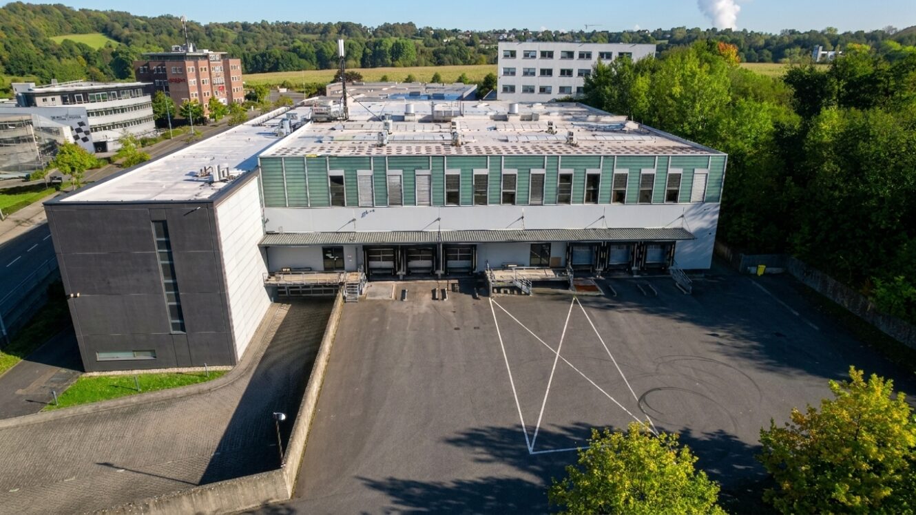 Ein großes Industriegebäude mit mehreren Laderampen und einem leeren, gepflasterten Vorplatz, umgeben von Bäumen und anderen Bürogebäuden.