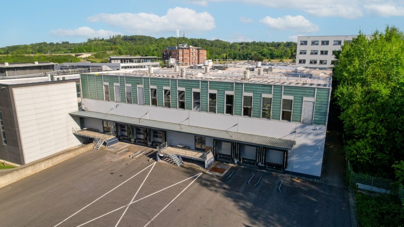 Ein großes, modernes Industriegebäude mit Laderampen ist von Parkplätzen und Bäumen umgeben, während der Himmel teilweise bewölkt ist.
