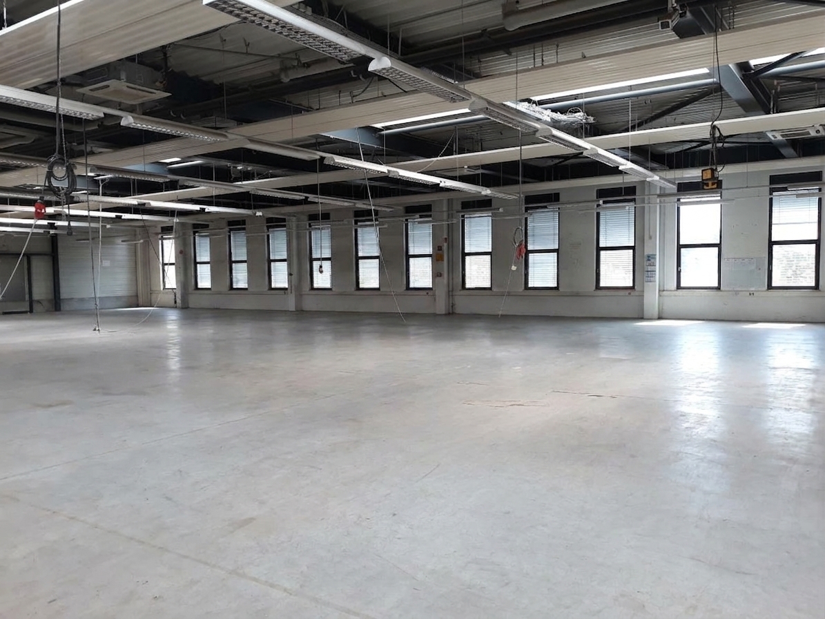 Großer, leerer Industrieraum mit weißen Böden, hohen Decken, freiliegenden Rohrleitungen und Fensterreihen entlang einer Wand.