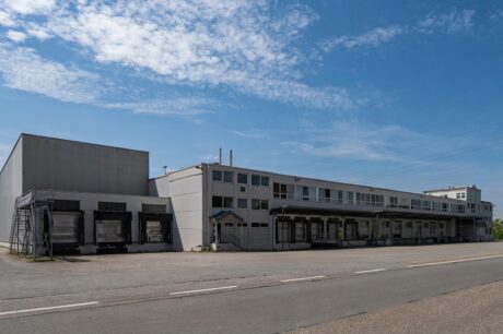 Große Industriehalle mit mehreren Laderampen und einer Außentreppe unter teilweise bewölktem Himmel.