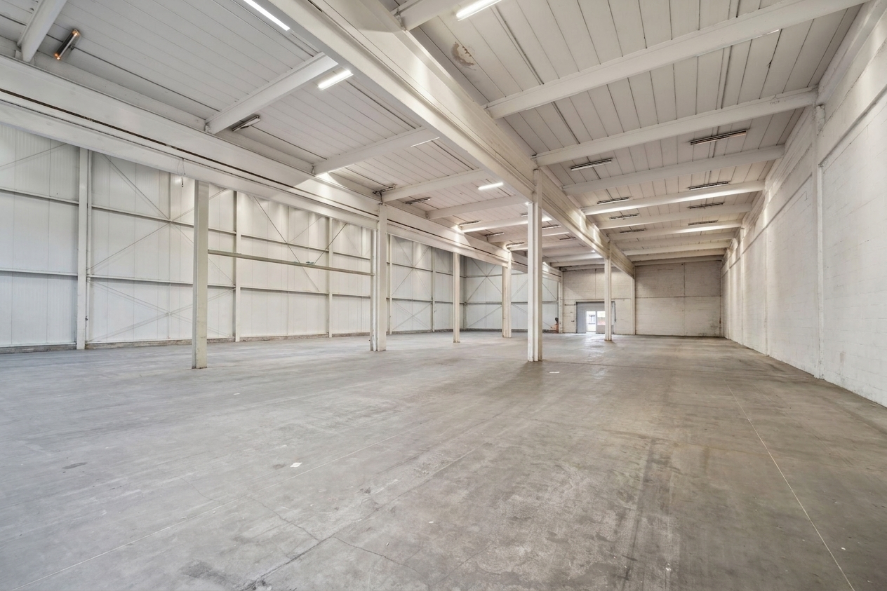 Leere Industriehalle mit hohen Decken, freiliegenden Balken, Betonboden und weißen Wänden, beleuchtet von Leuchtstoffröhren. Am anderen Ende ist eine kleine Tür zu sehen.