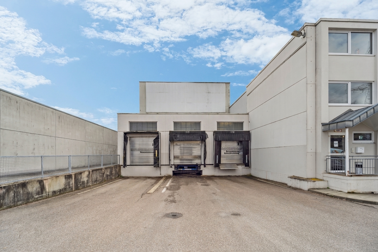 Zwei Laderampen mit geschlossenen Metallrollläden an einem schlichten Industriegebäude, umgeben von Betonmauern und unter einem größtenteils klaren Himmel.