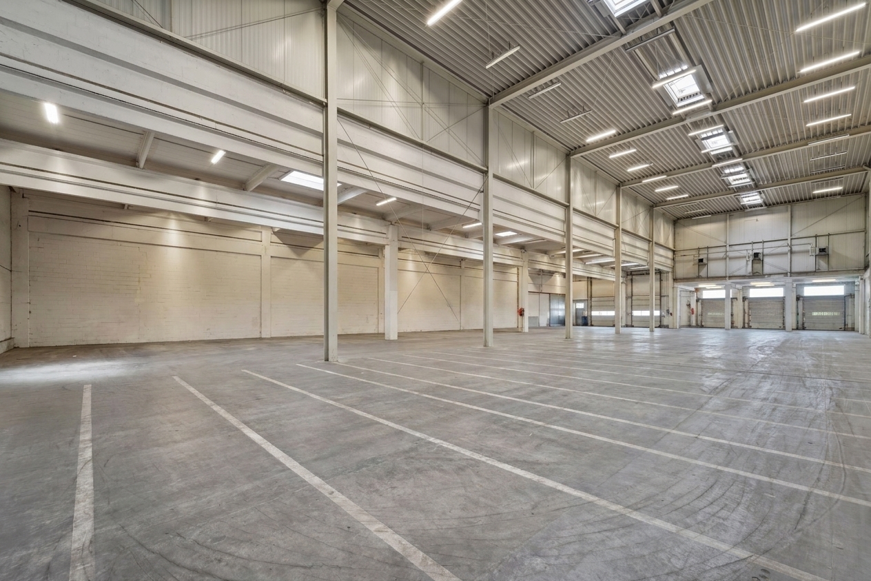 Große, leere Lagerhalle mit hohen Decken, freiliegenden Balken, Oberlichtern und markierten Park- oder Ladezonen auf dem Betonboden. Der Raum hat helle Wände und ist durch künstliche Beleuchtung hell ausgeleuchtet.