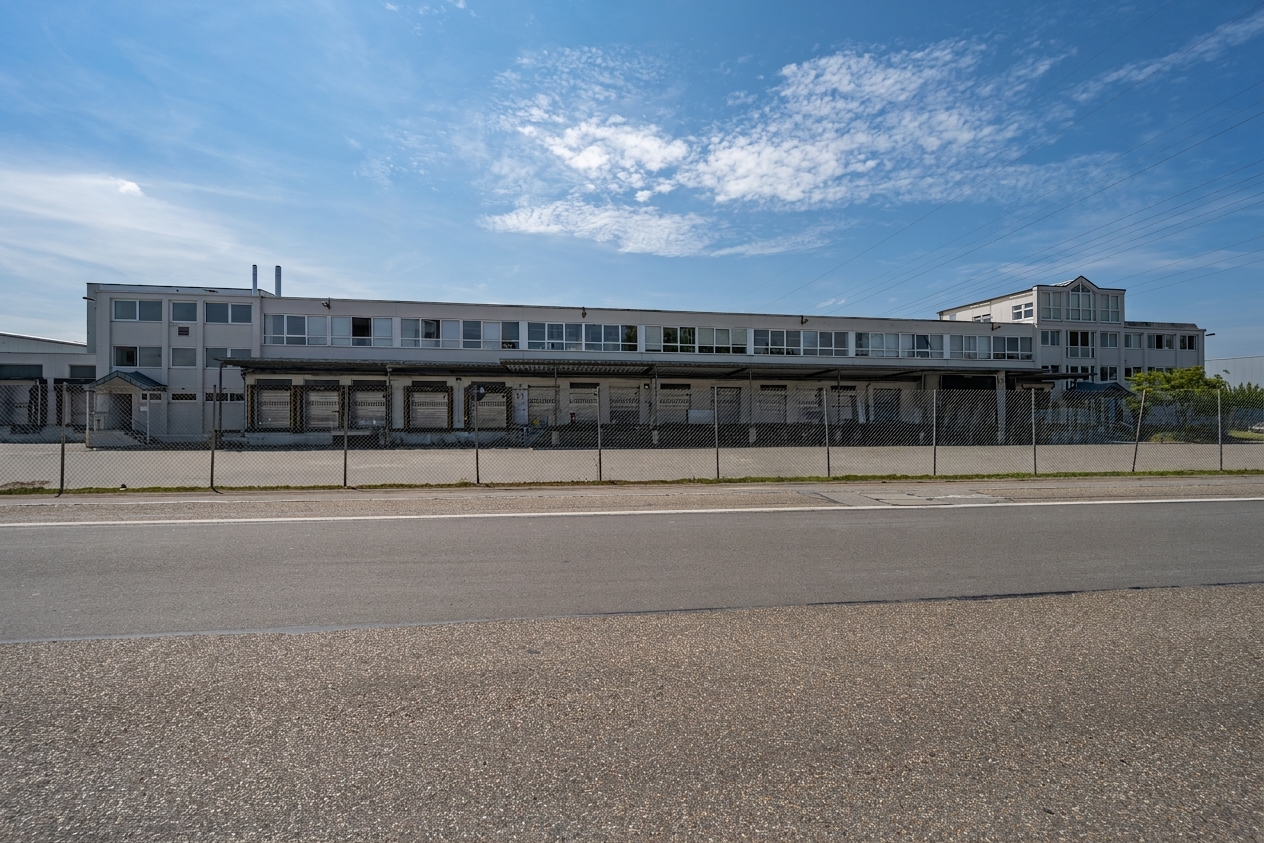 Ein langes, weißes Industriegebäude mit einem umzäunten Gelände steht neben einer leeren Straße unter einem blauen Himmel, der von vereinzelten Wolken übersät ist.