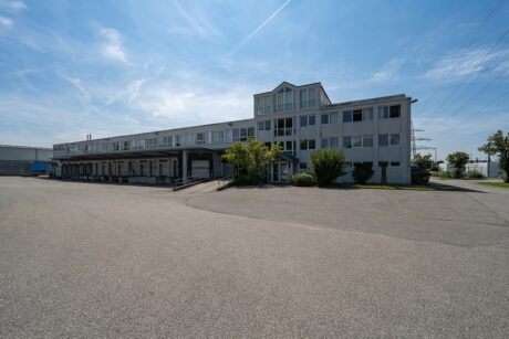 Unter blauem Himmel steht ein großes, graues Industriegebäude mit vielen Fenstern und einer Laderampe neben einem leeren, gepflasterten Gelände.
