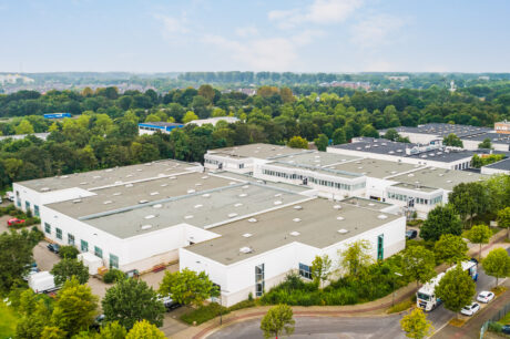 Luftaufnahme eines großen Industriekomplexes mit mehreren weißen Lagerhallen, umgeben von Bäumen und mehreren Lastwagen, die entlang einer angrenzenden Straße geparkt sind.