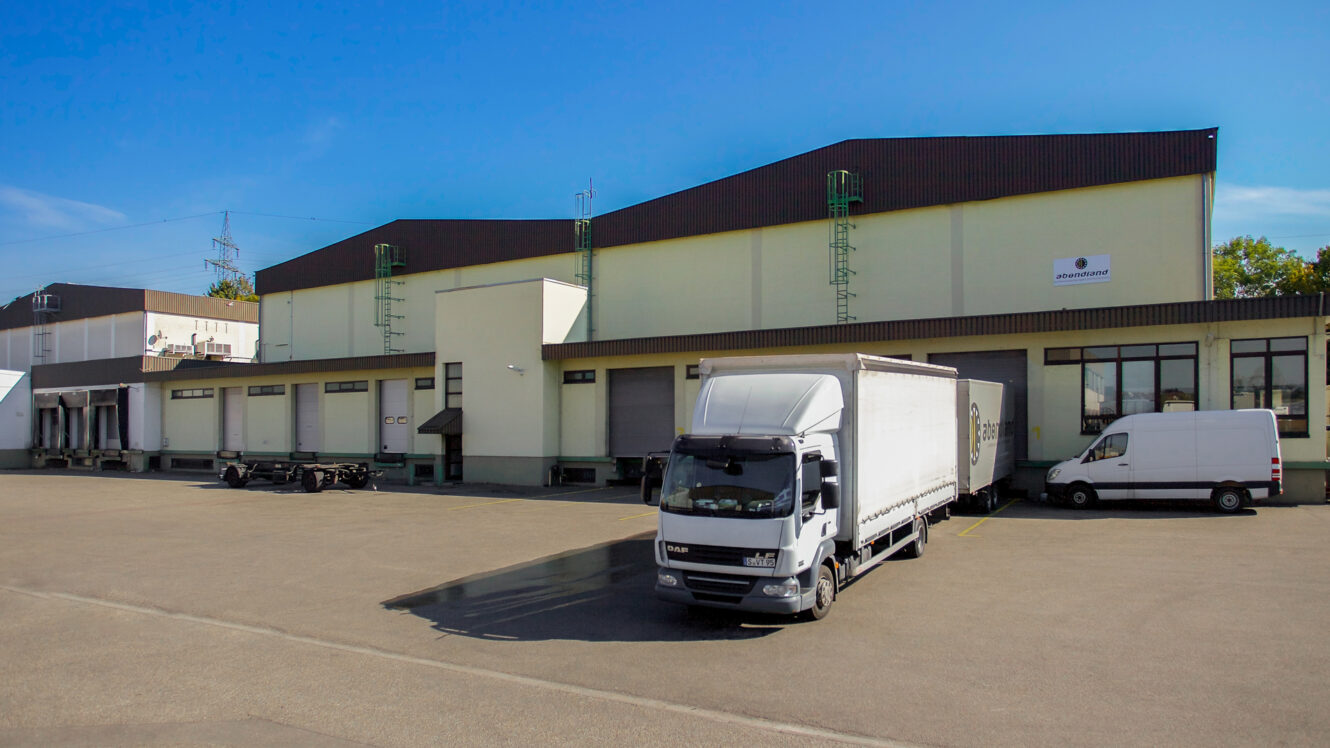 Ein weißer DAF-Lieferwagen, der vor einer großen Industriehalle mit mehreren Laderampen geparkt ist.