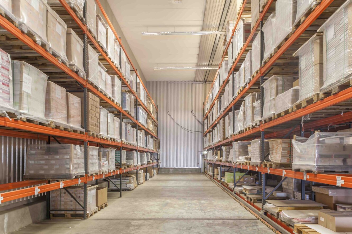 Reihen von Metallregalen, die in einem Lagergang hoch mit Paletten voller Kartons und in Schrumpffolie verpackter Waren gestapelt sind.