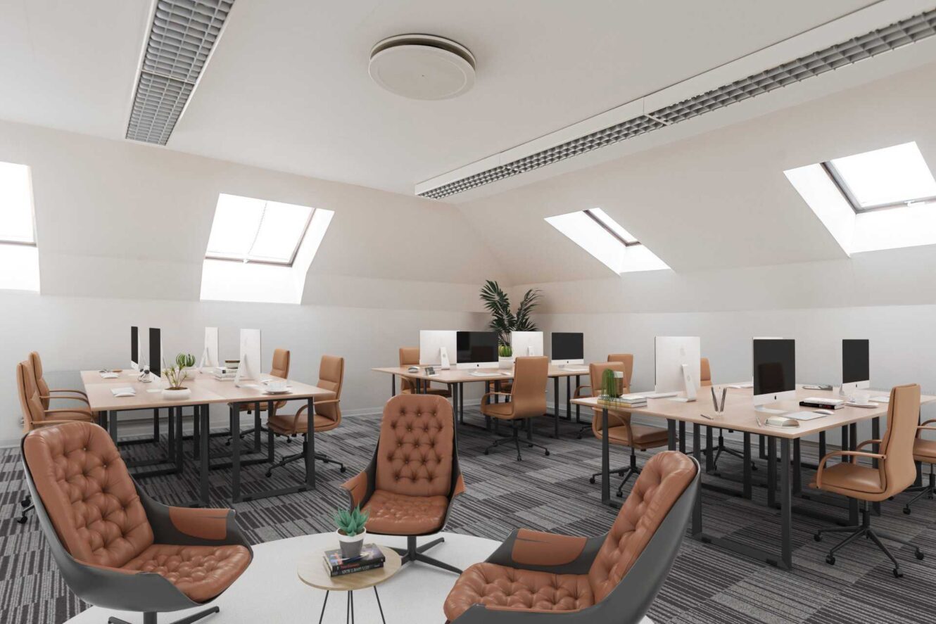 Modernt kontor i öppen planlösning med flera skrivbord, datorer, bruna stolar, takfönster och en liten sittgrupp med fåtöljer och ett runt bord.