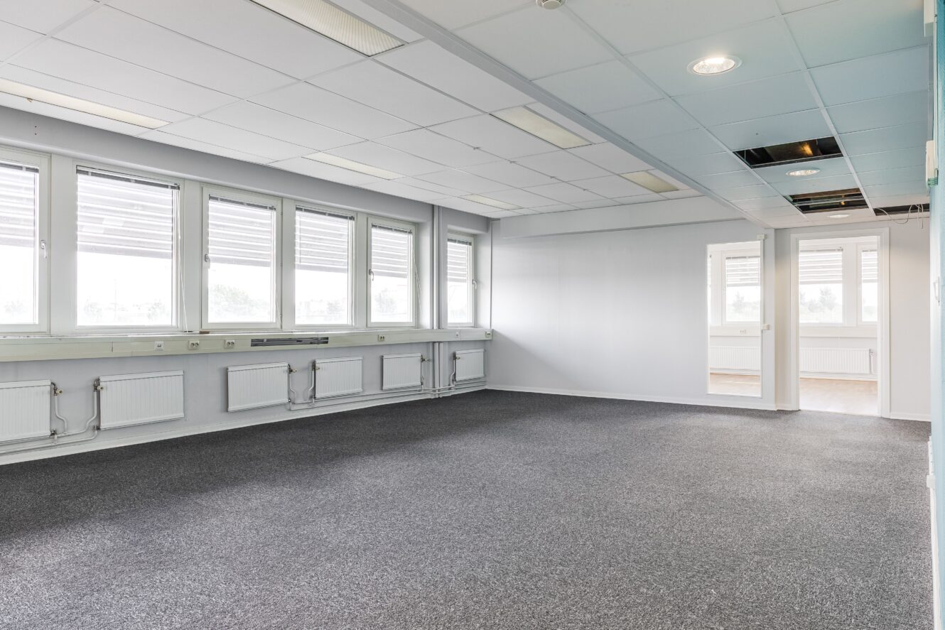 Tomt kontorsrum med grå heltäckningsmatta, flera stora fönster, vita väggar, taklampor och några takpaneler saknas.
