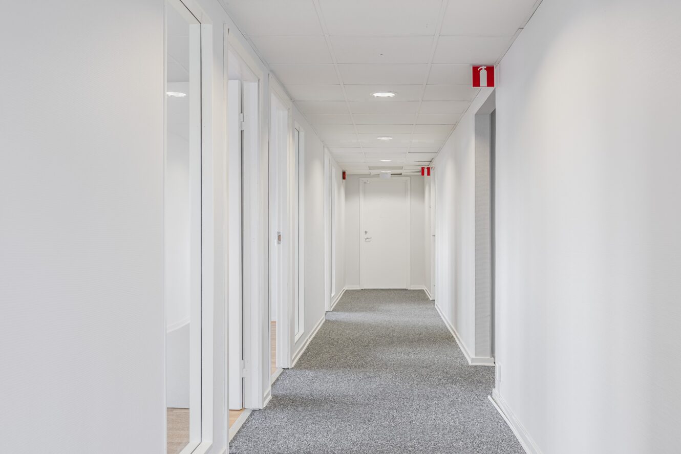 En ren, vit kontorskorridor med stängda dörrar på båda sidor, grå heltäckningsmatta, taklampor och röd brandsäkerhetsutrustning på väggarna.