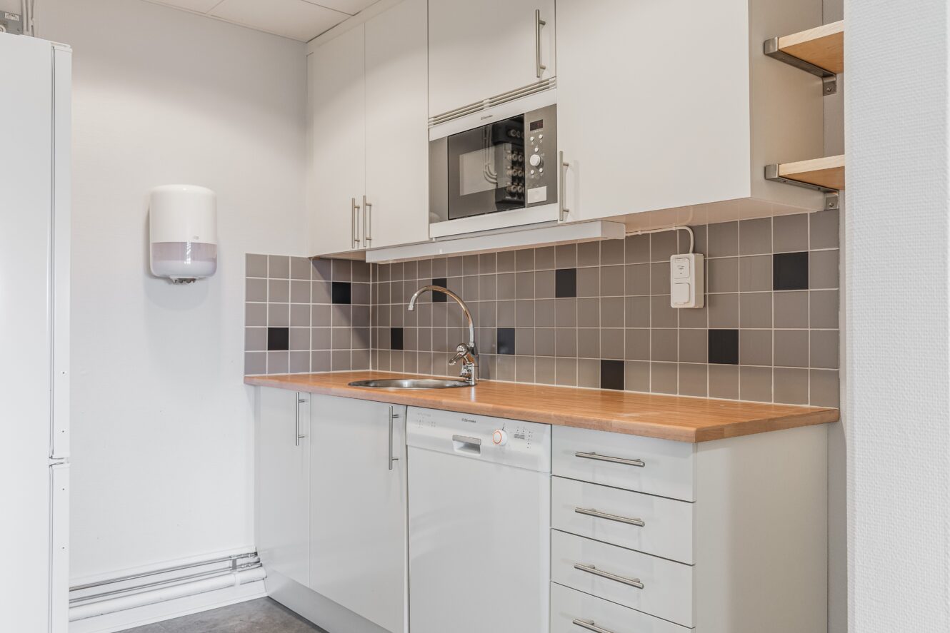 Modernt pentry med vita skåp, bänkskiva i trä, inbyggd mikrovågsugn, diskmaskin, diskho i rostfritt stål och kaklat stänkskydd med grå och svarta accenter.