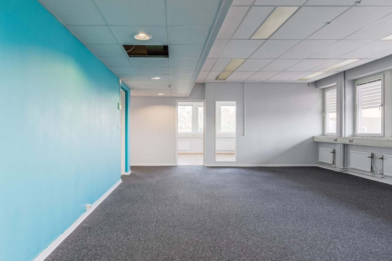 Tomt kontorsrum med blå accentvägg, grå heltäckningsmatta, vita takpaneler, lysrör och fönster längs ena sidan.