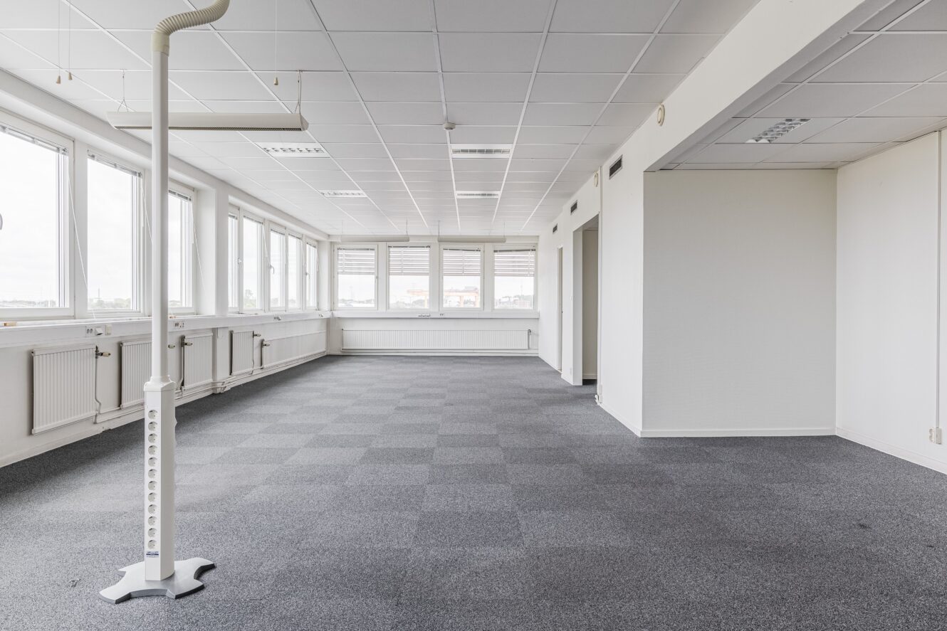 Tomt kontorsutrymme med grå matta, vita väggar, stora fönster längs ena sidan, taklampor och ett golvmonterat eluttag.