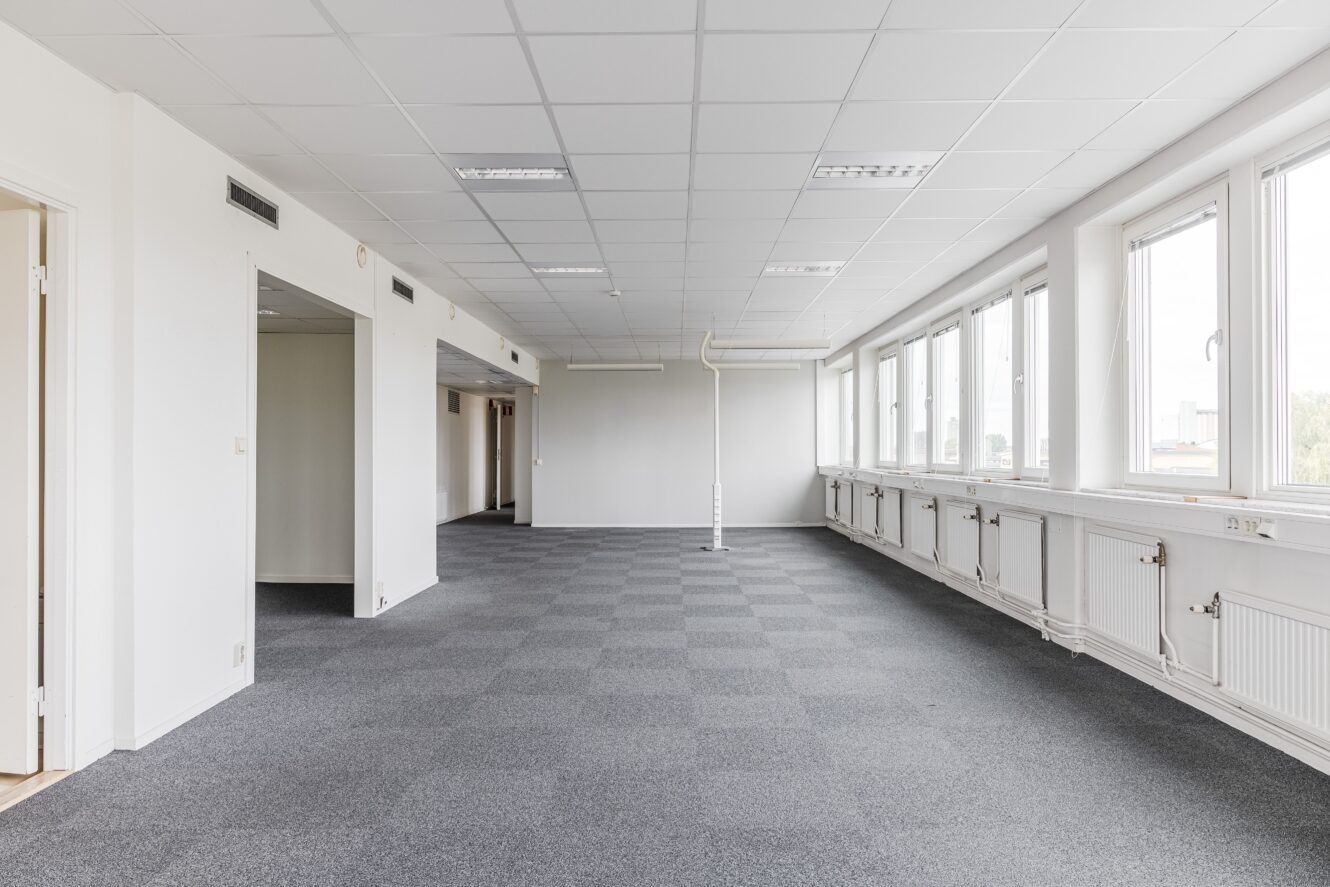 Tomt kontorsutrymme med grå heltäckningsmatta, vita väggar, taklampor, stora fönster längs ena sidan och flera öppna dörröppningar.