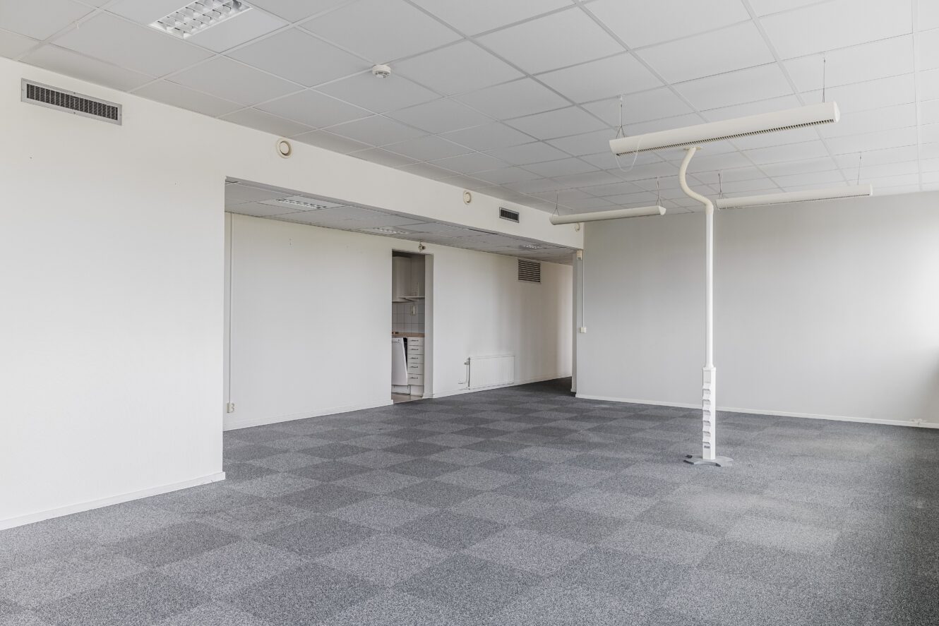 Tomt kontorsutrymme med grårutig matta, vita väggar, ett vitt tak med lysrör och en enda pelare i mitten.
