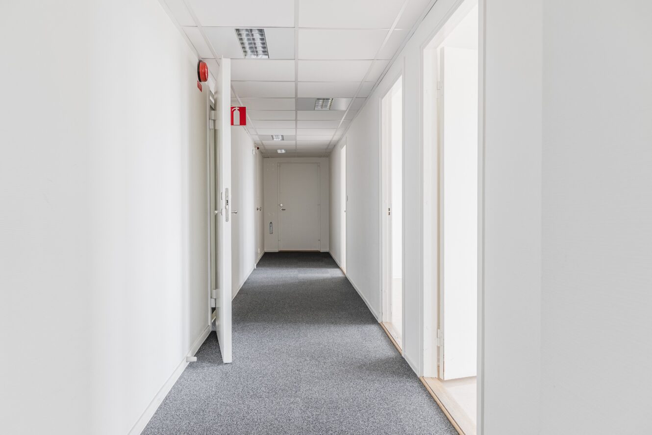 En ren, tom korridor med vita väggar, grå heltäckningsmatta, flera öppna dörrar och en stängd dörr i slutet. Lysrörslampor sitter i taket.