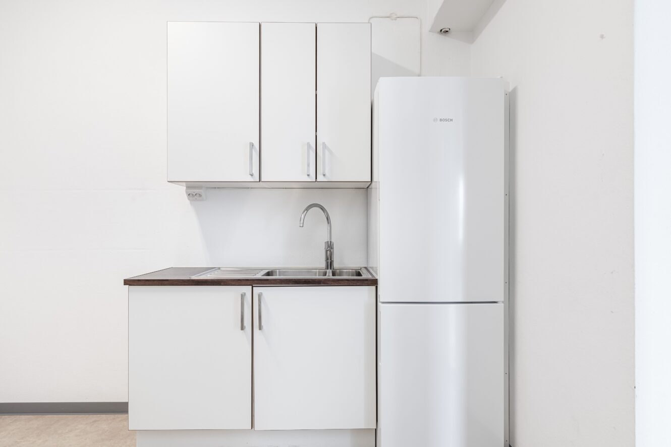 Litet kök med vita skåp, diskho i rostfritt stål, silverfärgad kran och ett vitt kylskåp mot en vit vägg.