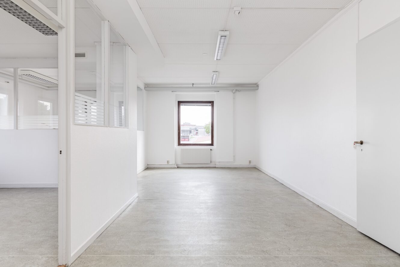 Tomt vitt kontorsrum med fönster, glaspartier till vänster, vita väggar, taklampor och en stängd dörr till höger.