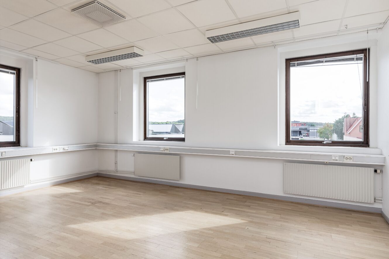 Tomt, omöblerat rum med ljust trägolv, vita väggar, tre stora fönster, taklampor och synliga radiatorer under fönstren.