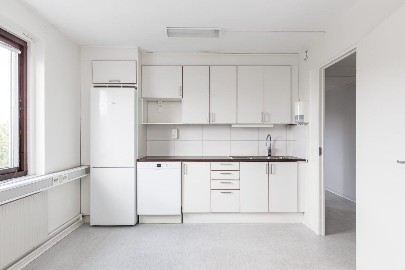 Minimalistiskt vitt kök med över- och underskåp, kylskåp, diskmaskin, diskho och ett fönster på vänster sida. Utrymmet ser rent och tomt ut.