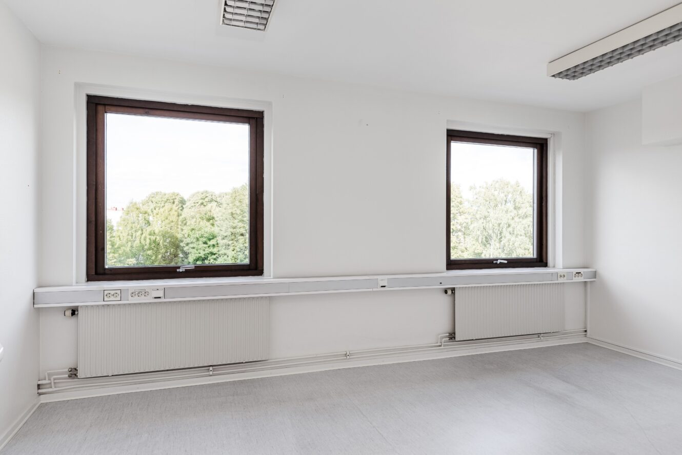 Tomt vitt rum med två stora fönster, element under varje fönster och utsikt över gröna träd utanför.