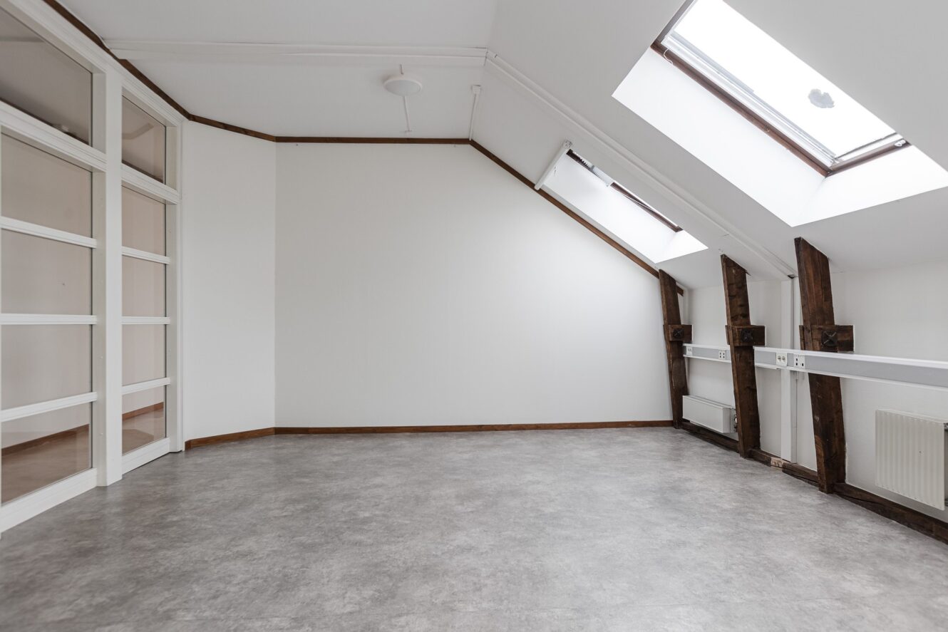 Tomt vindsrum med snedtak, stora takfönster, synliga bjälkar, grått golv, vita väggar och en glasad skiljevägg.